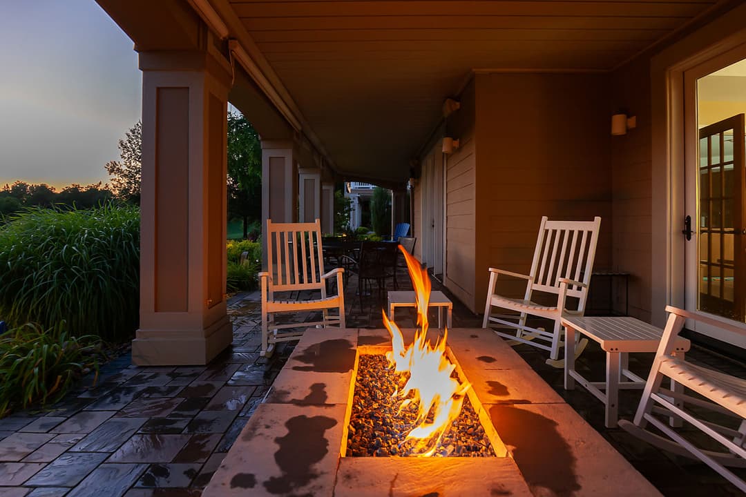 Cozy patio with a fire pit, stone seating, and rocking chairs at dusk, surrounded by greenery.
