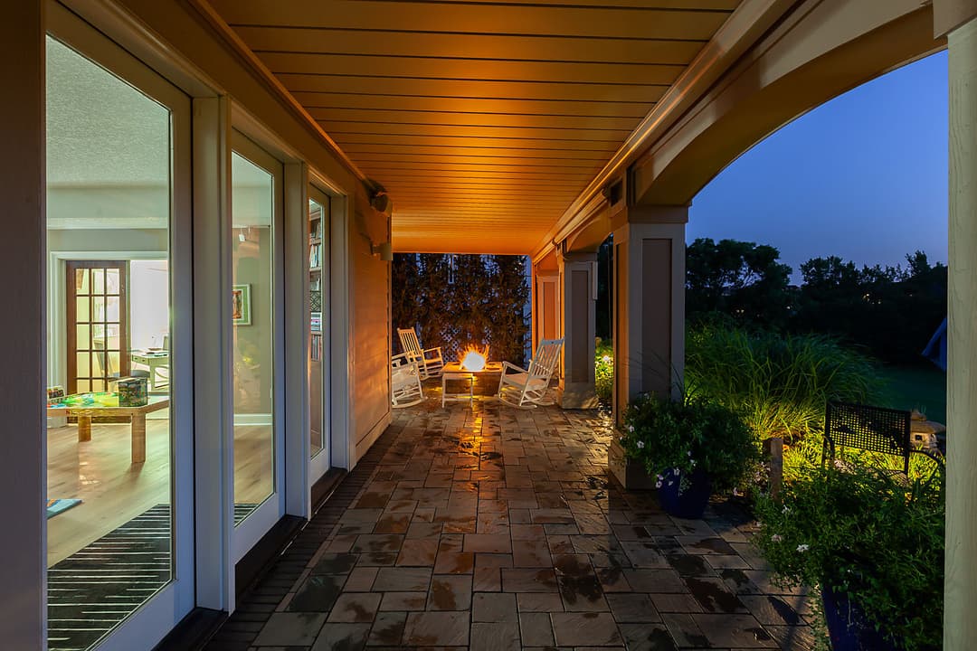 Cozy porch with stone flooring, glowing fire pit, and illuminated greenery at dusk.