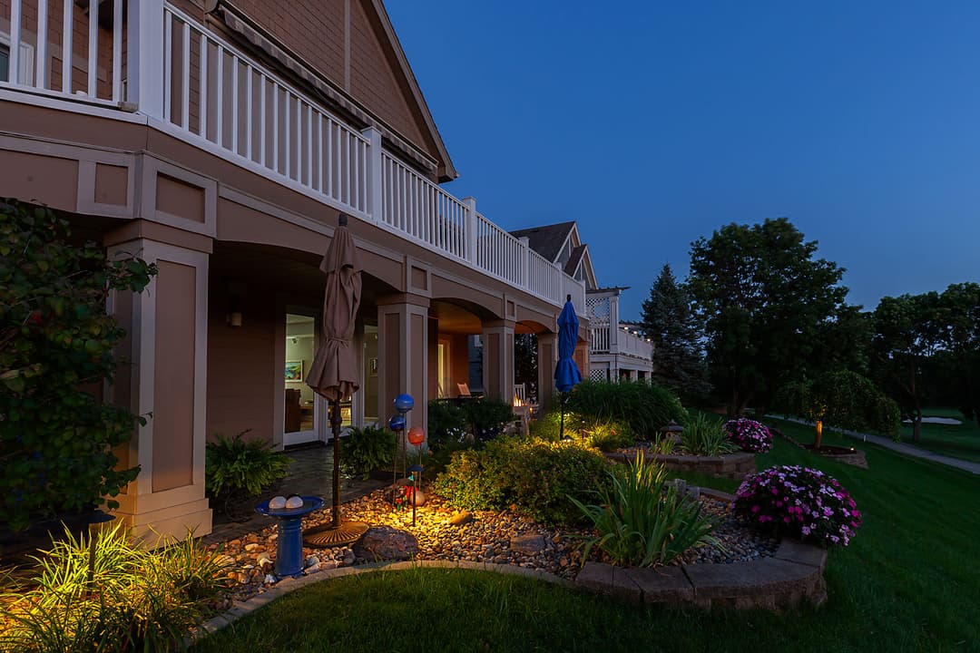 Illuminated landscape of a home exterior at dusk with flowers and outdoor seating.