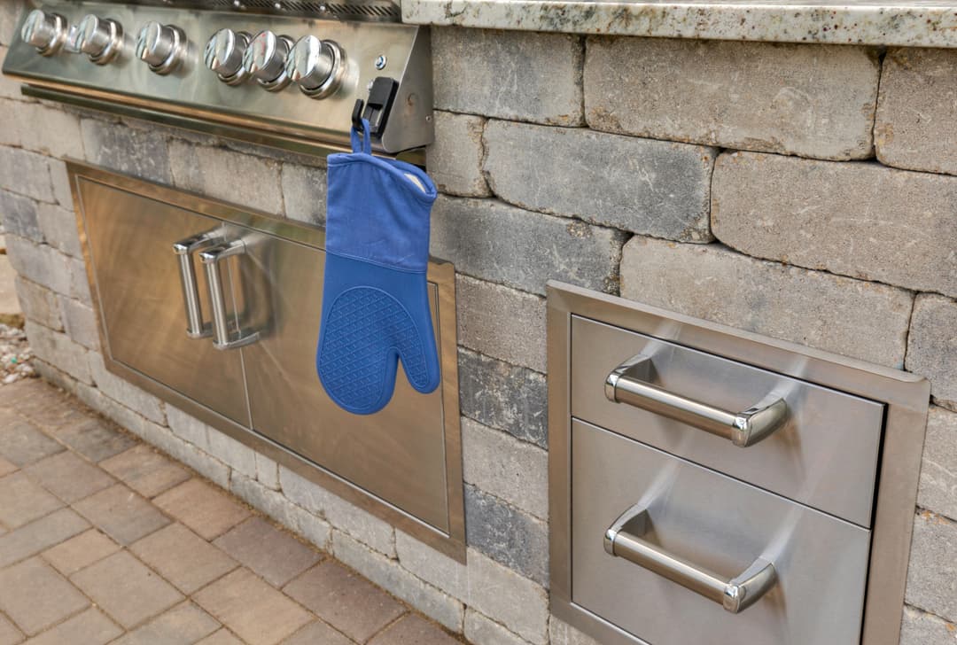 Outdoor kitchen features stainless steel grill, stone countertops, and blue oven mitt hanging.