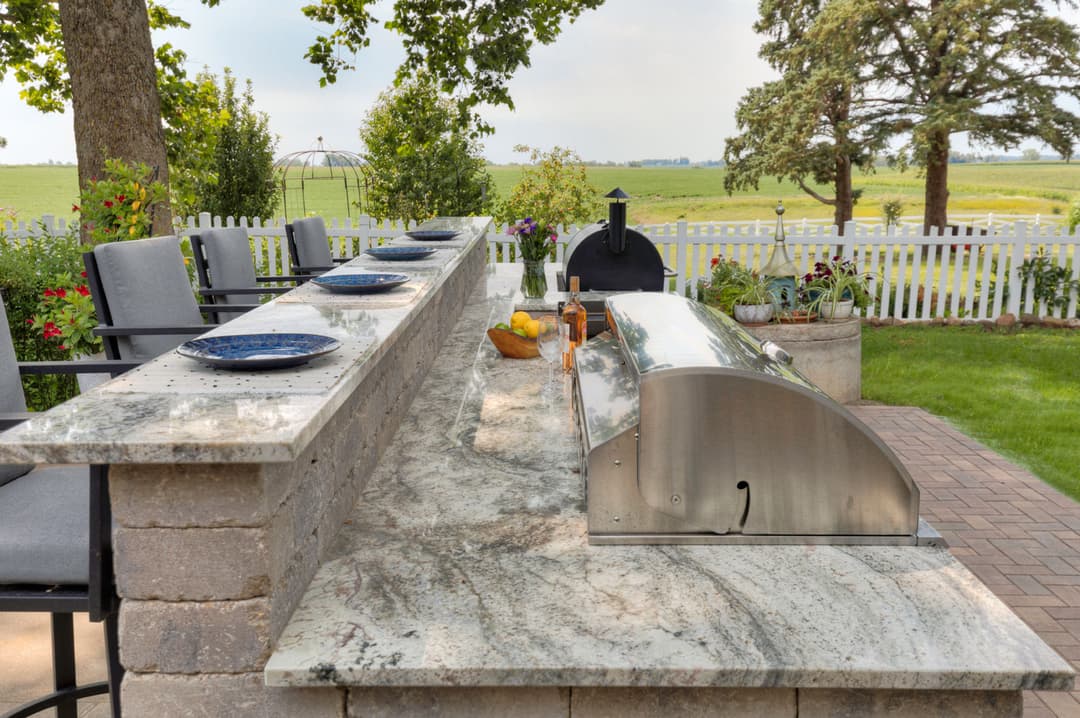 Outdoor kitchen with granite countertop, BBQ grill, and dining setup amidst green landscape.