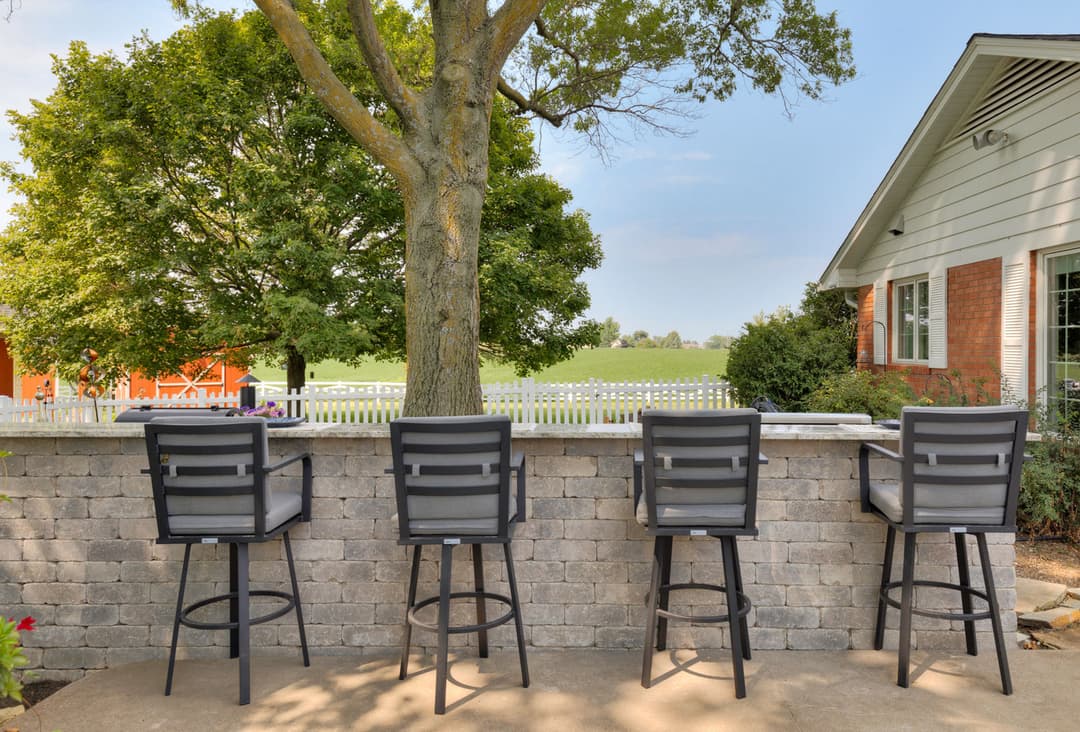Outdoor seating area with four barstools at a stone wall overlooking a green field.
