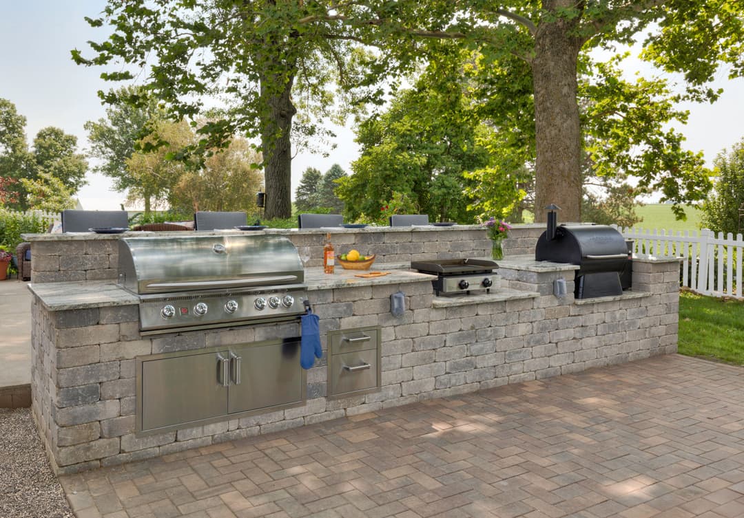 Outdoor kitchen setup with stainless steel grill and smoker, surrounded by lush greenery.