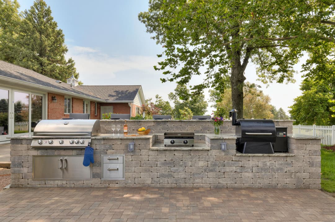 Outdoor kitchen featuring a stainless steel grill, smoker, and brick counter in a backyard setting.