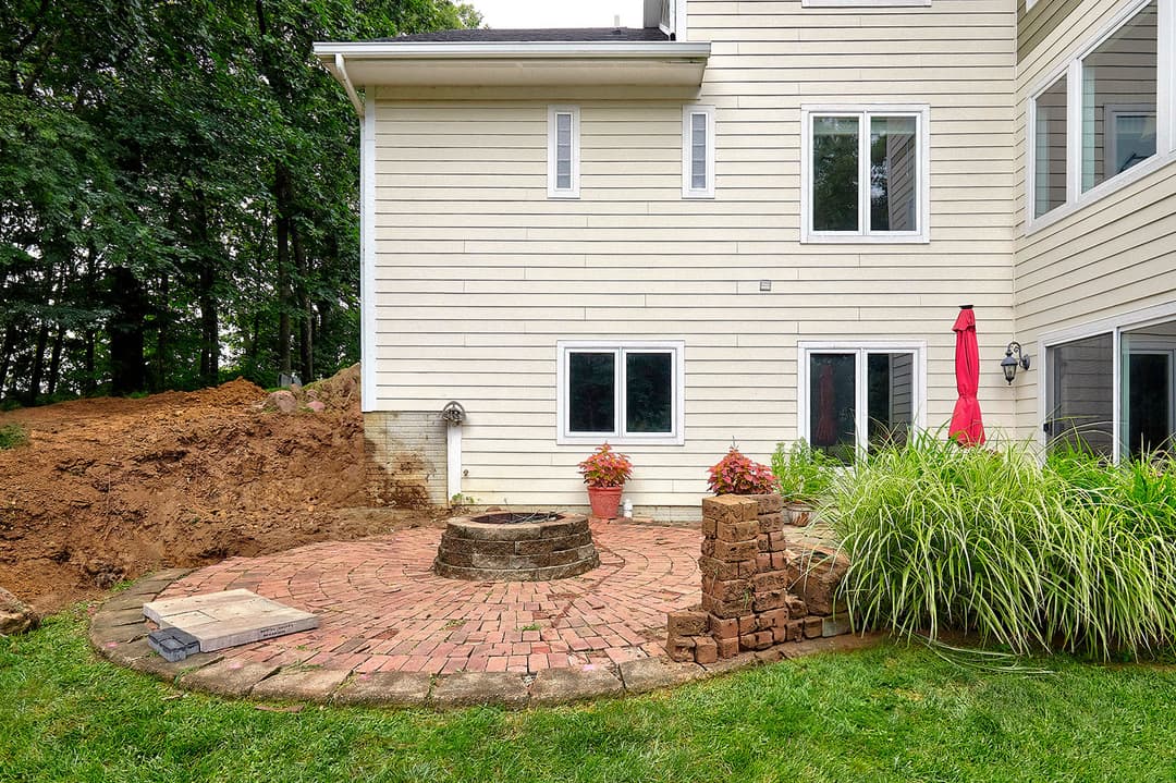 Patio with brick fire pit and landscaping near a house, surrounded by greenery and trees.
