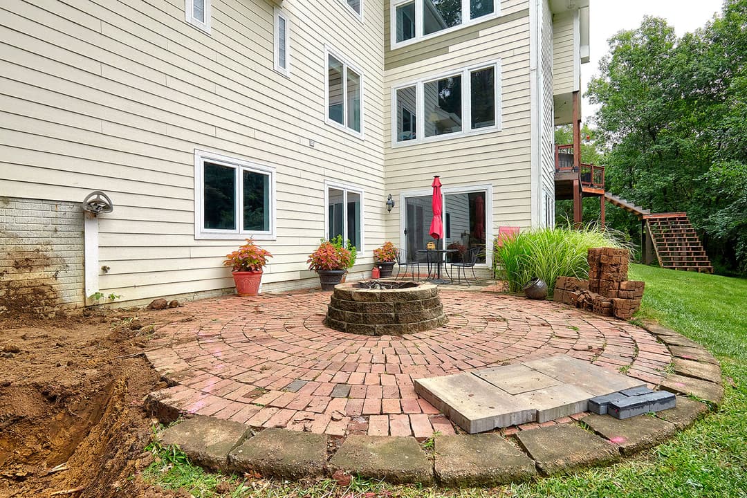 Patio with brick circular fire pit and green lawn, surrounded by plants and a house exterior.