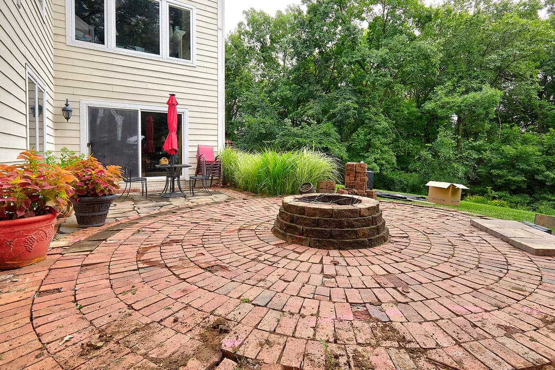 Backyard patio with circular brick design, potted plants, and wooded backdrop.
