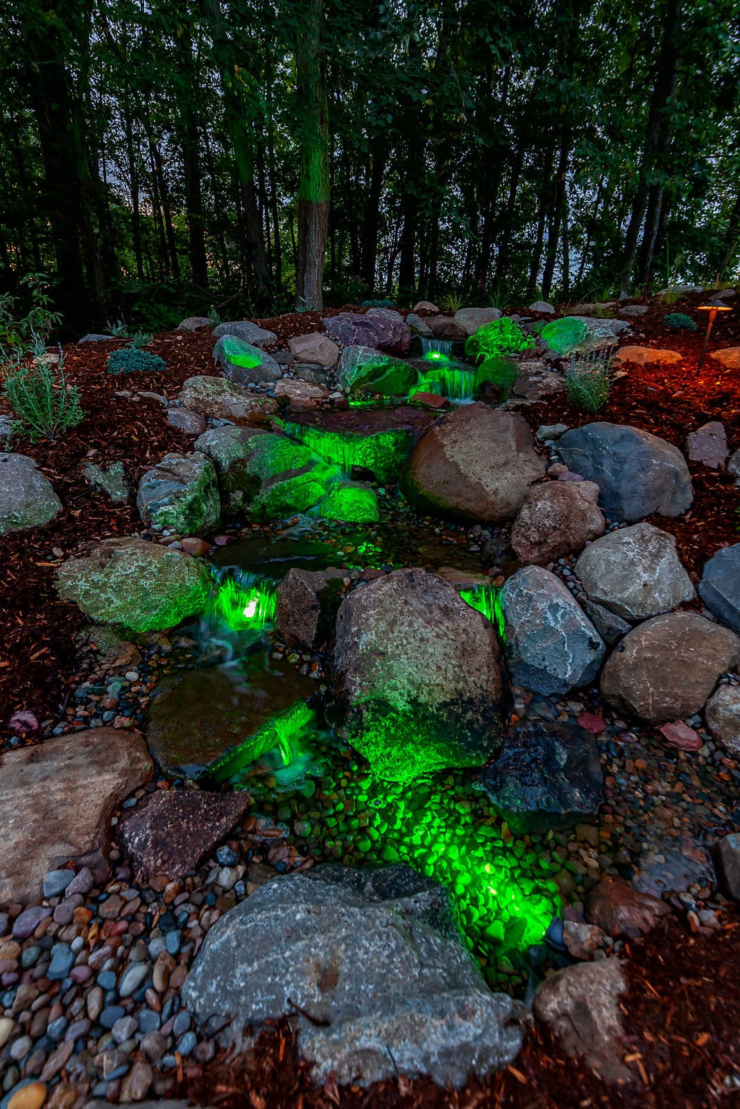 Illuminated stone landscape with green lights and a flowing water feature in a forest setting.