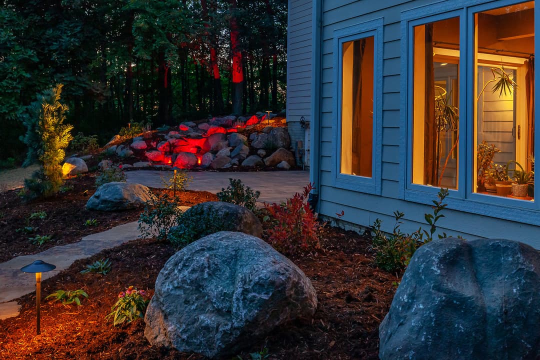 Landscape at dusk featuring a home's exterior with illuminated garden rocks and plants.