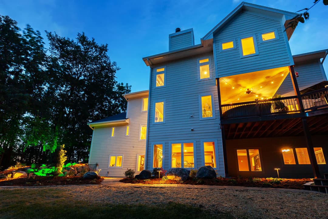 Modern home exterior at dusk with illuminated windows and landscaped yard featuring rocks and greenery.