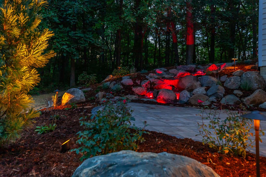 Illuminated rock garden with red lights, surrounded by shrubs and trees at dusk.