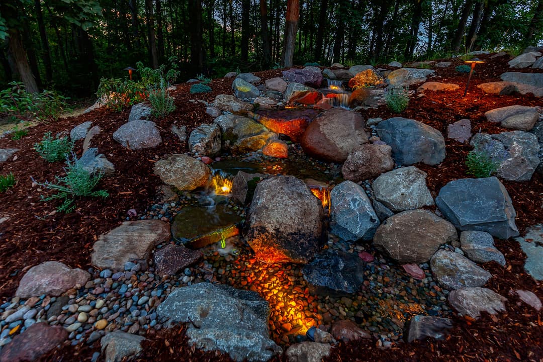 Serene garden water feature with rocks, illuminated by colorful lights among lush greenery.