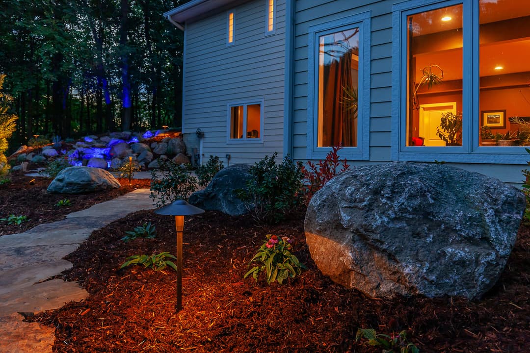 Cozy home exterior with pathway, garden lighting, and lush landscaping at twilight.