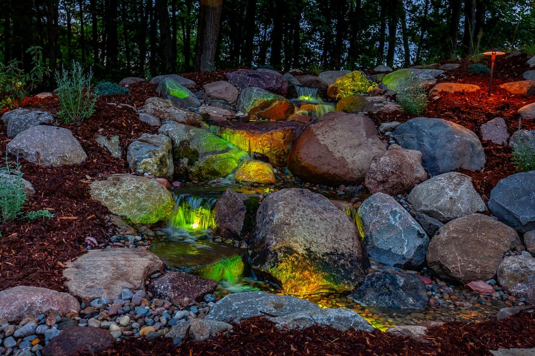 Serene stone garden waterfall with colorful lights and lush landscaping at dusk.