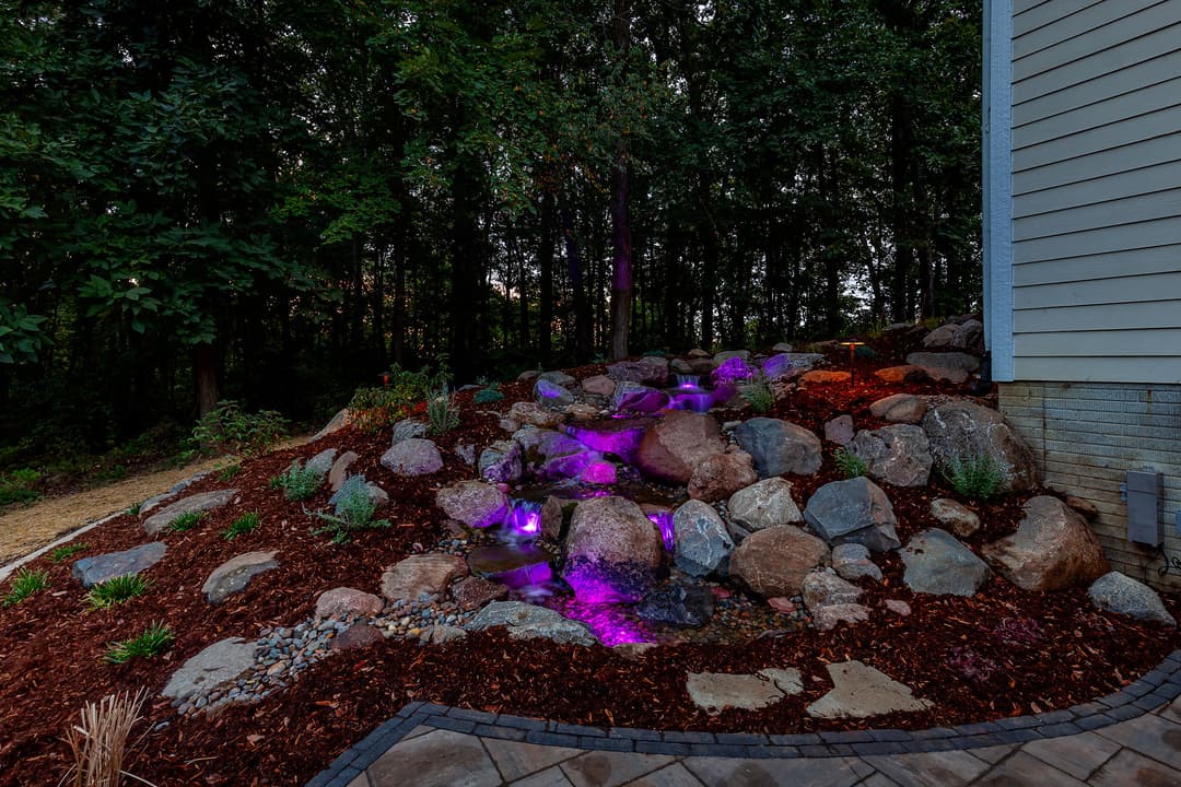 Illuminated rock waterfall feature in a landscaped garden with lush greenery.