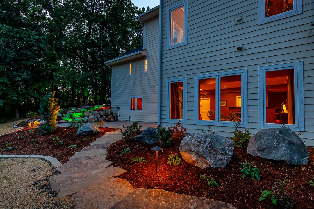 Brightly lit home exterior with landscaped garden, seating area, and decorative rocks at twilight.