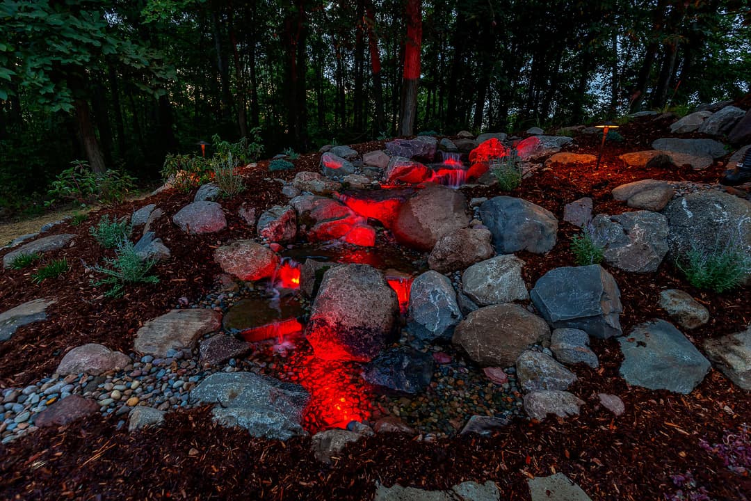 Illuminated rock garden waterfall with red lights, surrounded by lush greenery and mulch.