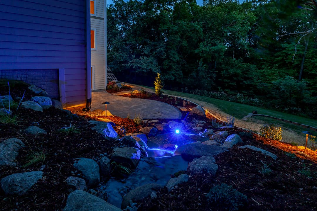 Backyard landscape at night featuring a illuminated water feature and stone pathways.