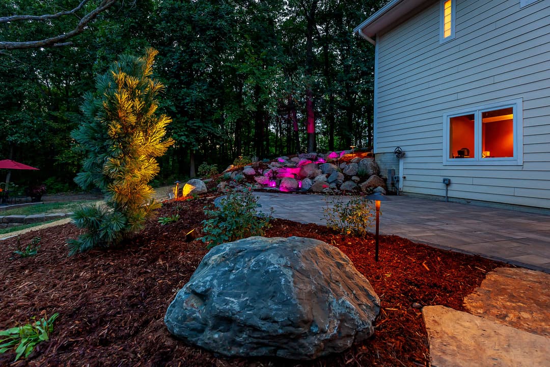 Illuminated backyard landscape featuring stone patio, greenery, and decorative boulders.