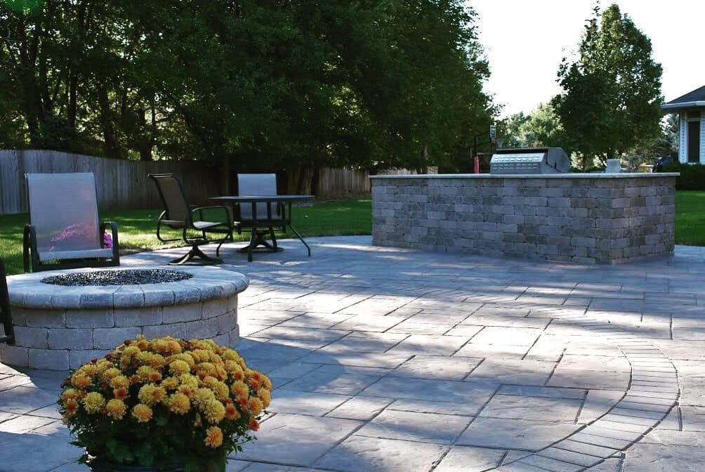 Patio featuring a stone fire pit, dining table, and outdoor grill surrounded by greenery.