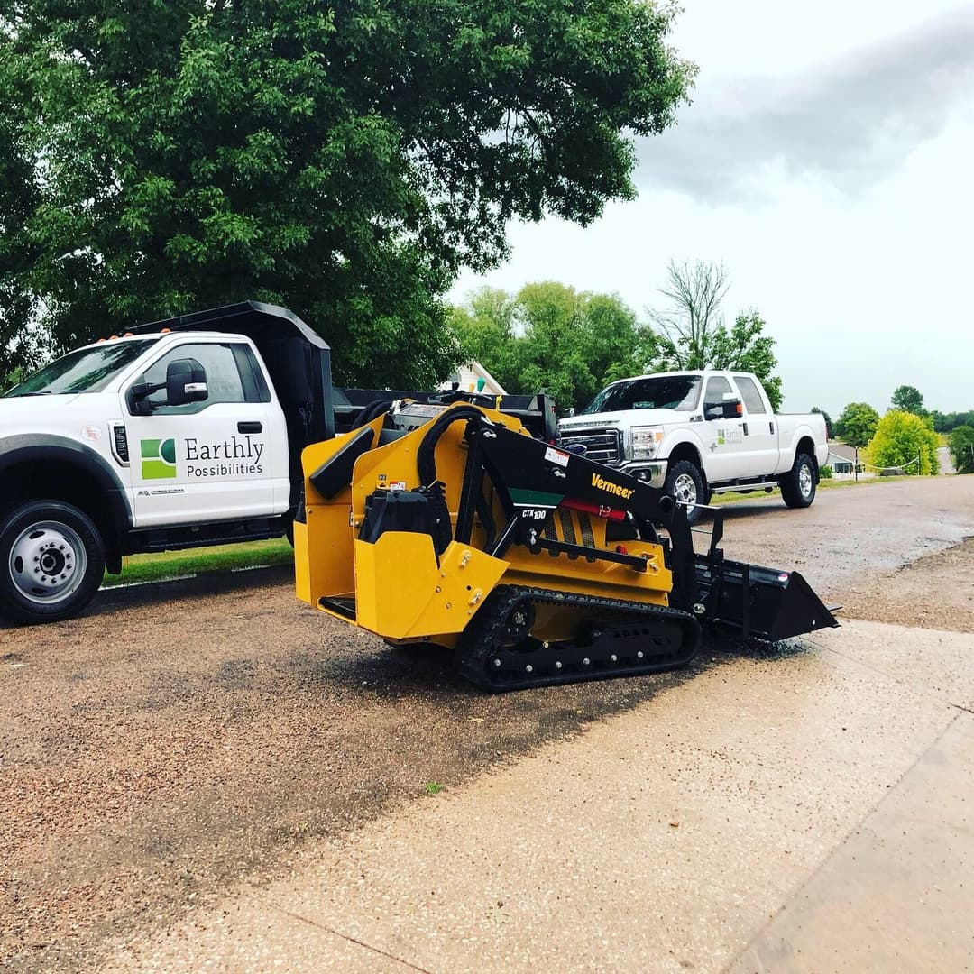 Compact track loader parked beside a truck labeled 'Earthly Possibilities' on a gravel road.