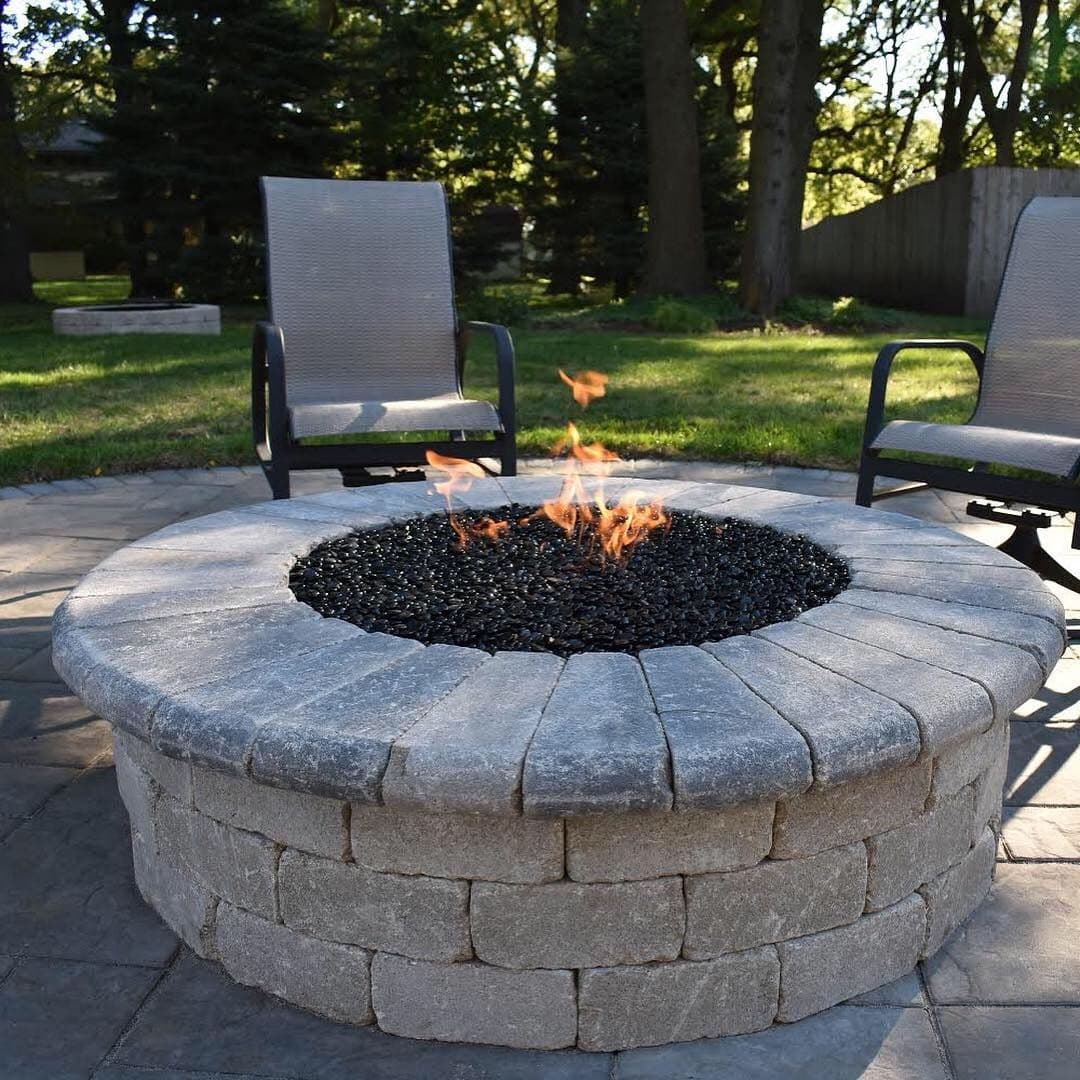 Modern circular fire pit with black glass stones, surrounded by outdoor chairs in a lush garden.