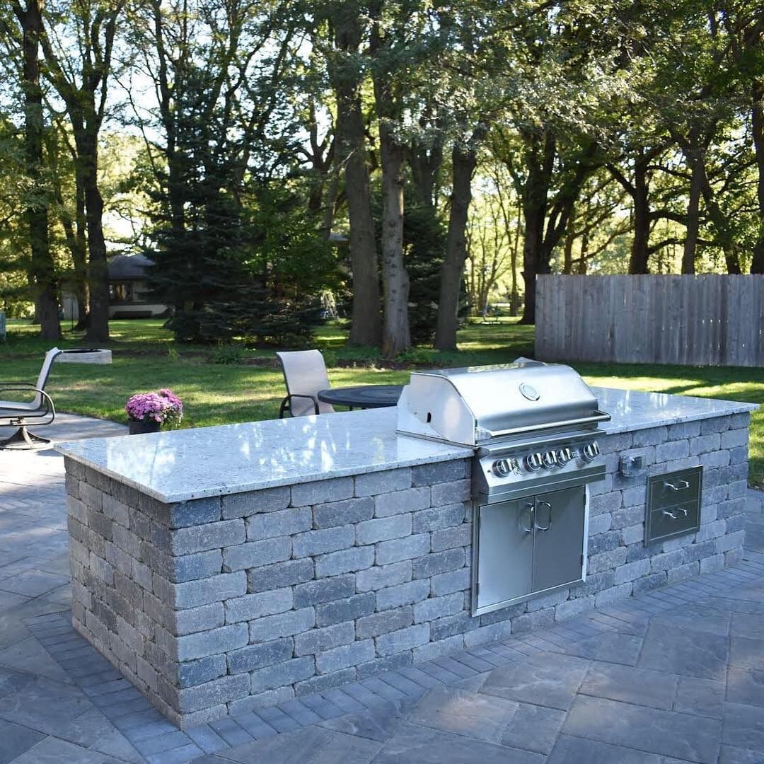 Stylish outdoor kitchen with stone grill island in a lush backyard setting.