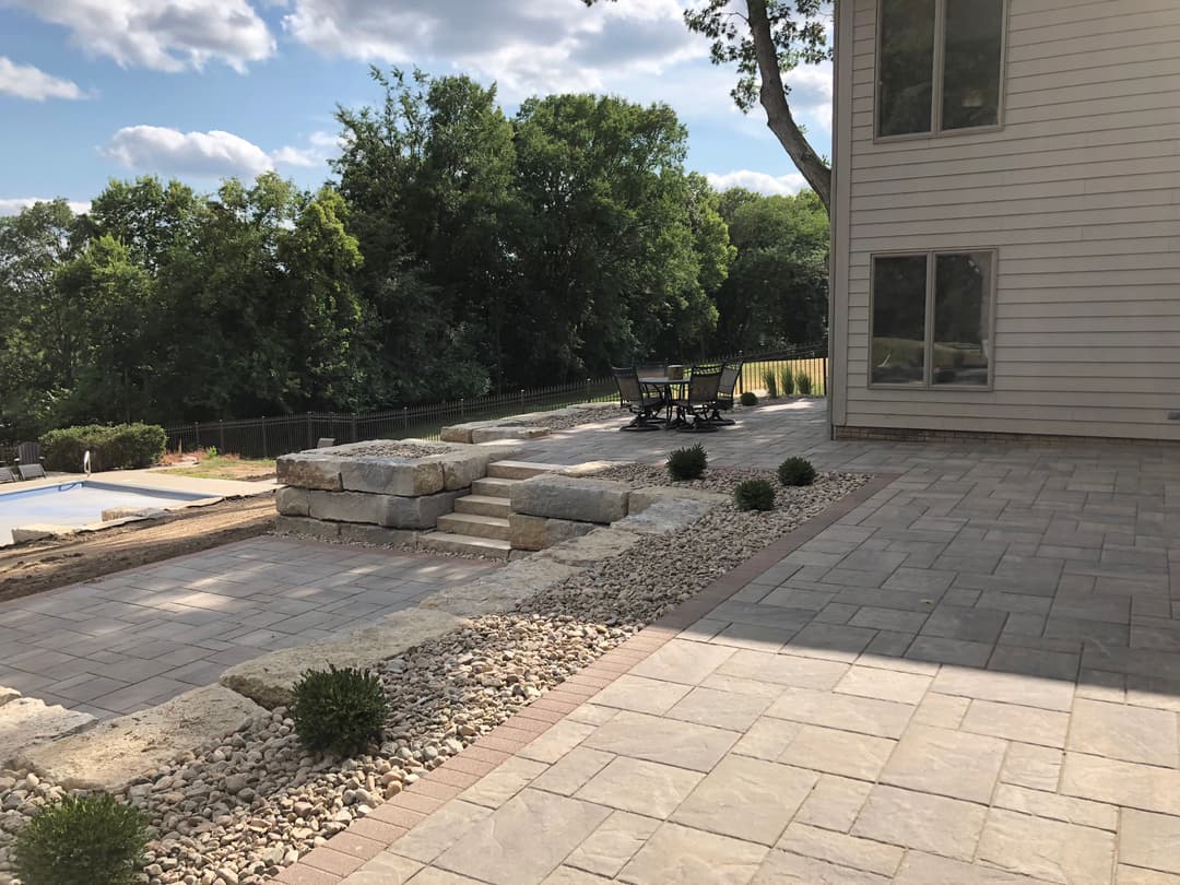 Patio area with stone steps, seating, and landscaped greenery under a clear blue sky.