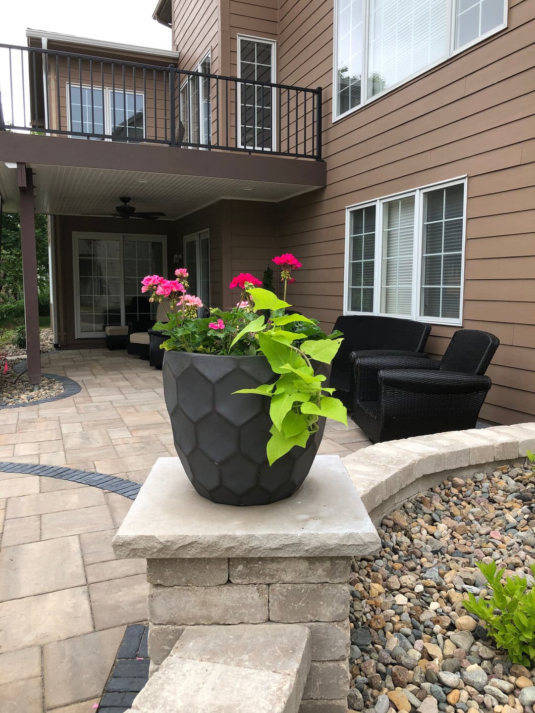 Black hexagonal planter with pink flowers on a patio near a house with large windows.