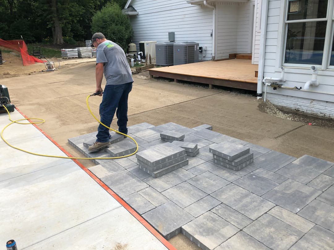 Worker laying gray pavers in outdoor patio area with tools and machinery nearby.