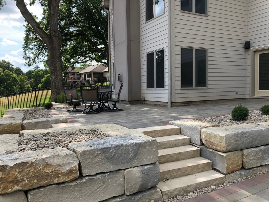 Patio area with stone steps, gravel, and seating near a home and green landscape.