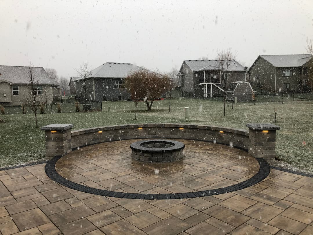 Snowy backyard with a stone fire pit and patio, surrounded by houses and falling snowflakes.