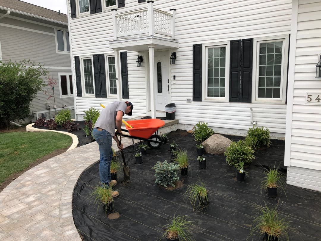 Man landscaping yard with plants, using a wheelbarrow and digging tools near a white house.