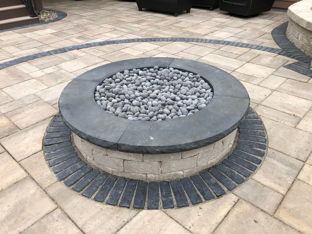 Modern circular fire pit with gray stones, surrounded by patterned stone pavers. Outdoor setting.