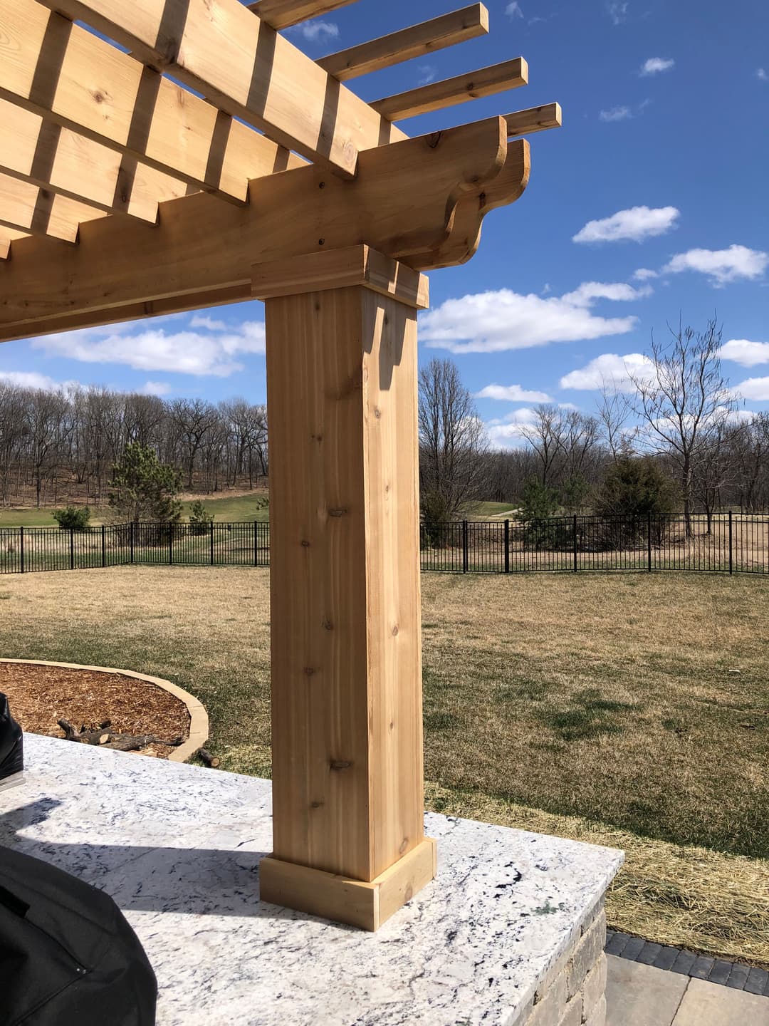 Wooden pergola post against a clear sky and landscaped yard, showcasing detailed craftsmanship.