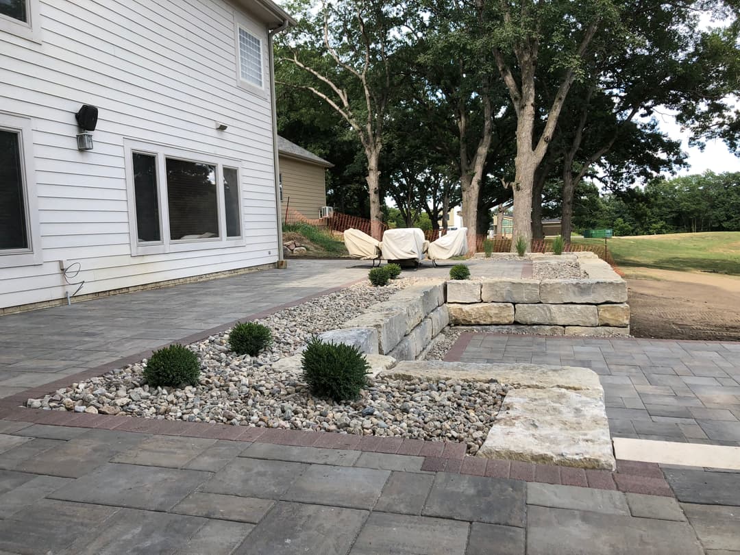 Patio with stone wall, gravel landscaping, and outdoor seating beside a house and trees.