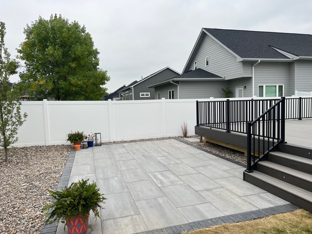 Modern backyard patio with gray pavers, black railing deck, and green landscaping.