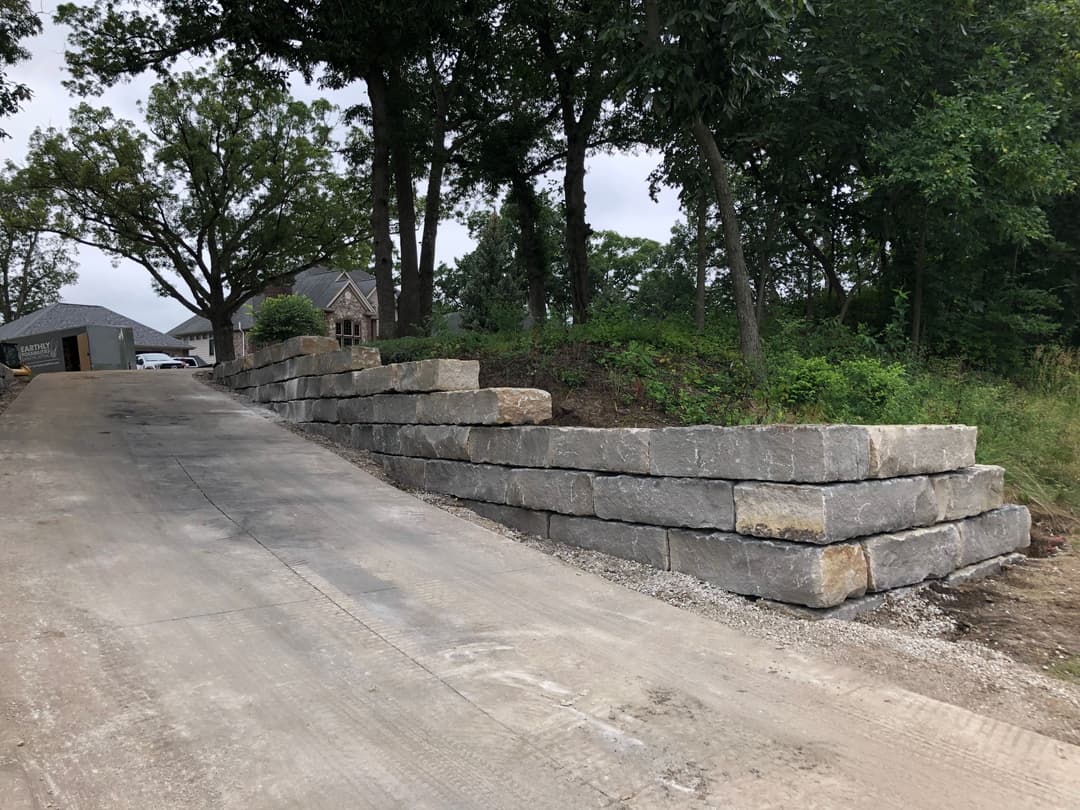 Stone retaining wall landscaping alongside a gravel driveway, surrounded by trees.