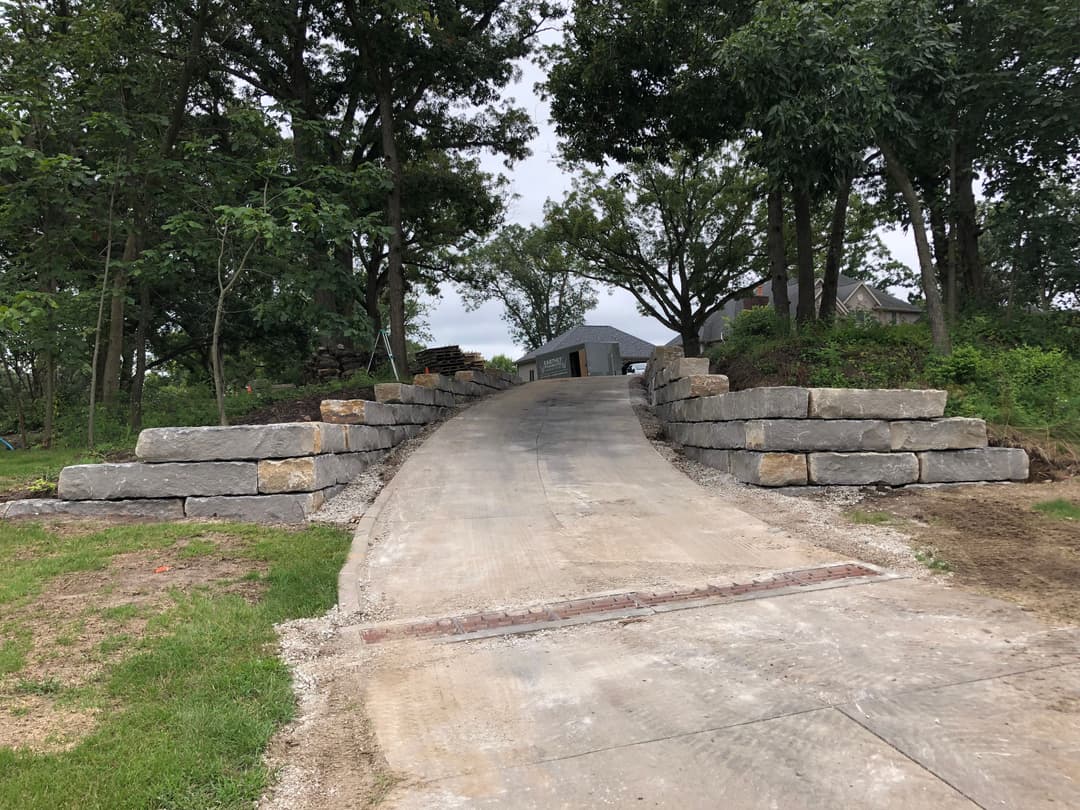 Driveway leading to a house, bordered by large stone retaining walls and surrounded by greenery.