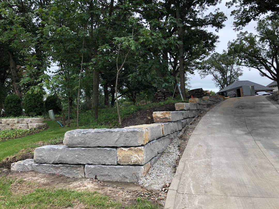 Stone retaining wall next to a winding driveway in a landscaped yard with trees.