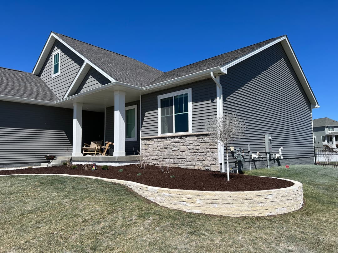 Newly landscaped home exterior with stone accents and a lush garden bed under a clear blue sky.