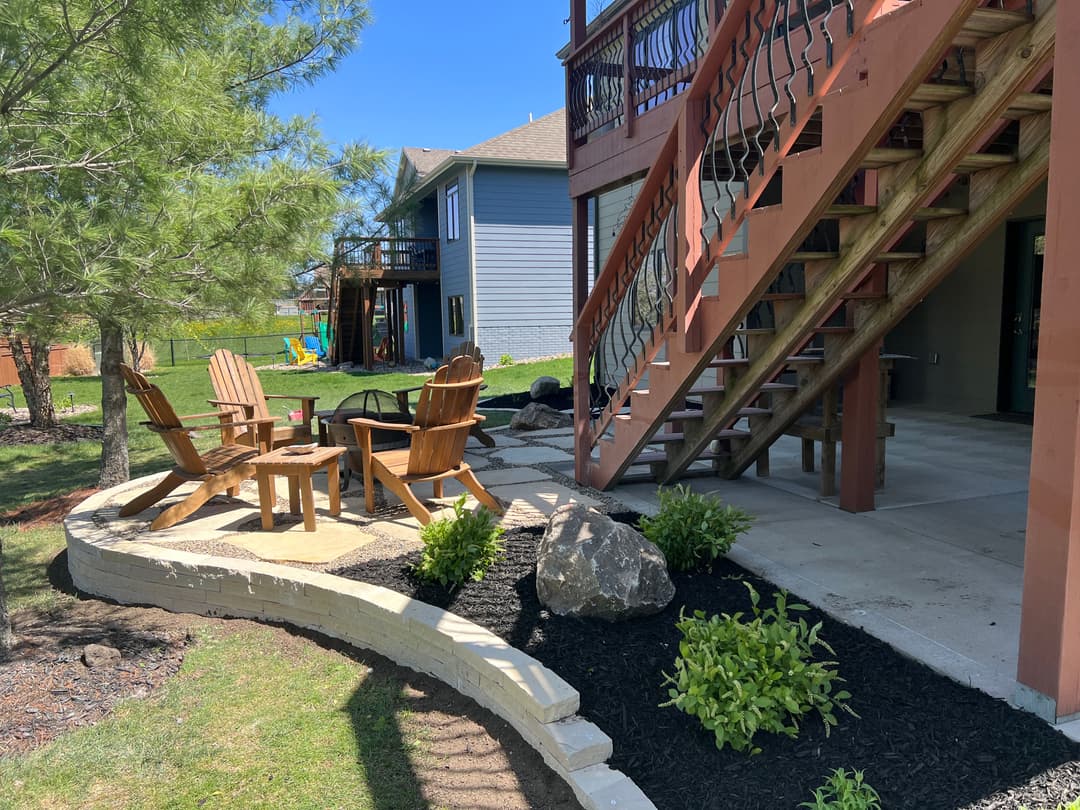 Backyard patio with Adirondack chairs, fire pit, and landscaped greenery under a sunny sky.