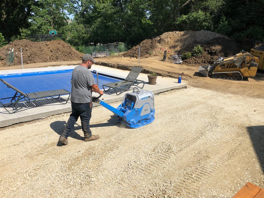 Worker operating a vibratory plate compactor on gravel near pool construction site.