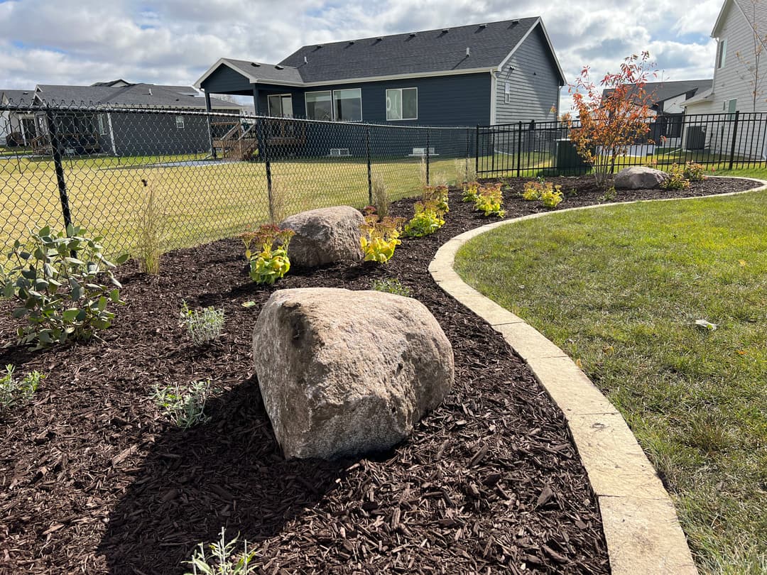 Landscaped garden with mulch, rocks, and plants near a fence and houses under a cloudy sky.
