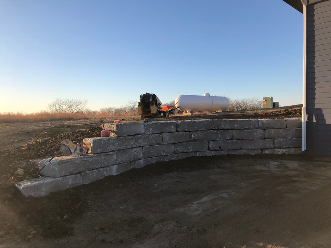 Excavator near a stone retaining wall and propane tank at a construction site at sunset.