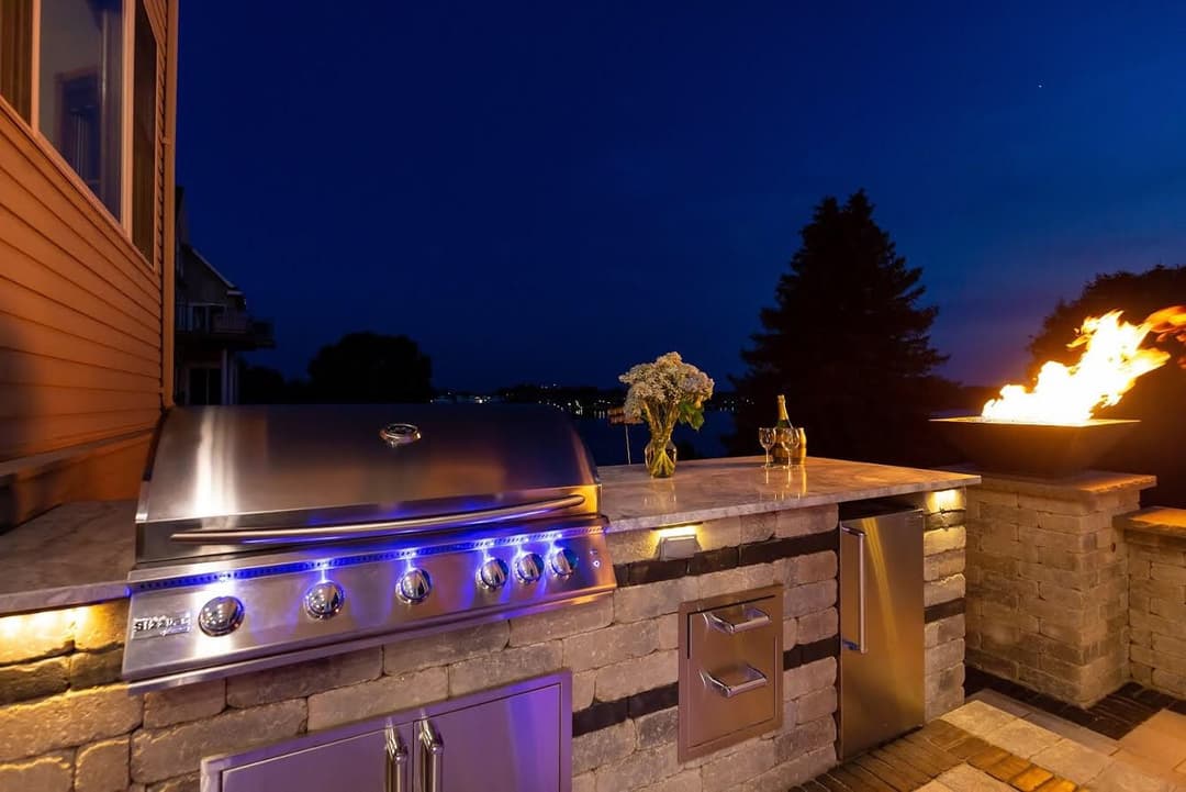 Modern outdoor kitchen with a stainless steel grill, stone countertop, and fire pit at night.