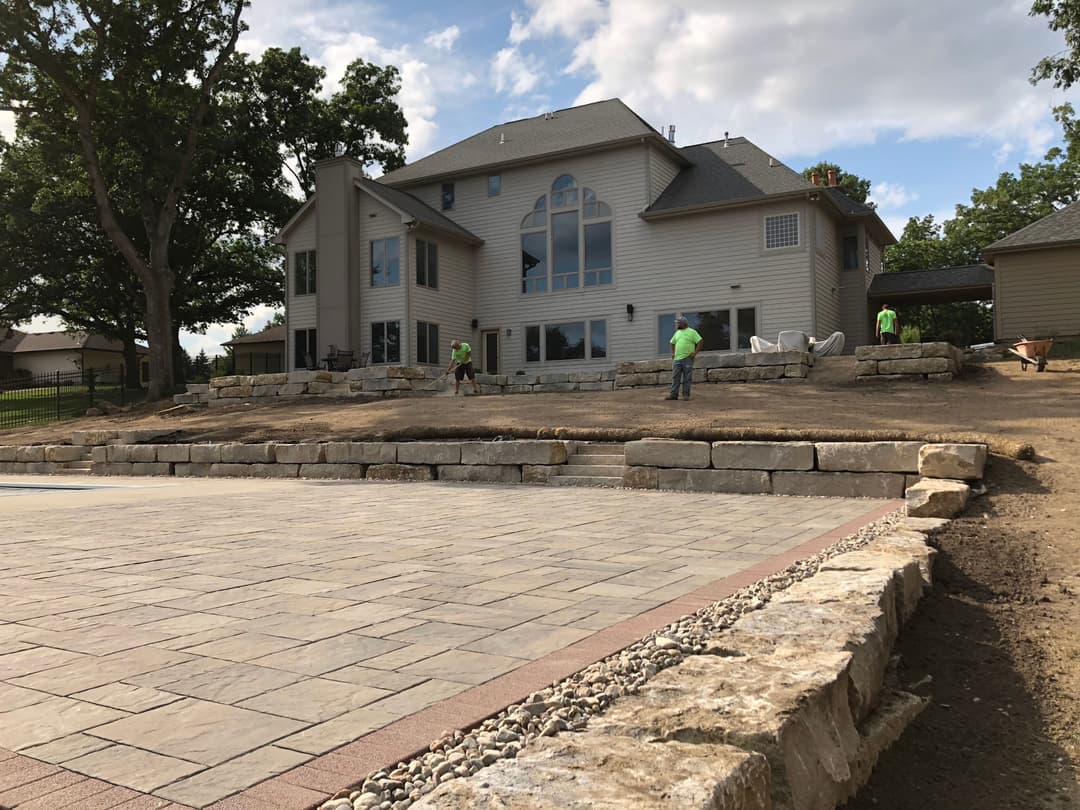 Landscape renovation with stone retaining wall near modern house and workers in green shirts.