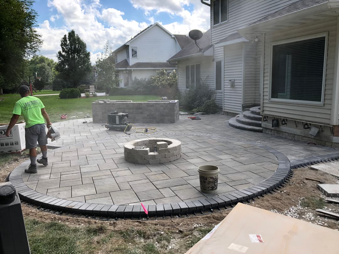 Worker installing a paver patio with a circular fire pit in a residential backyard.