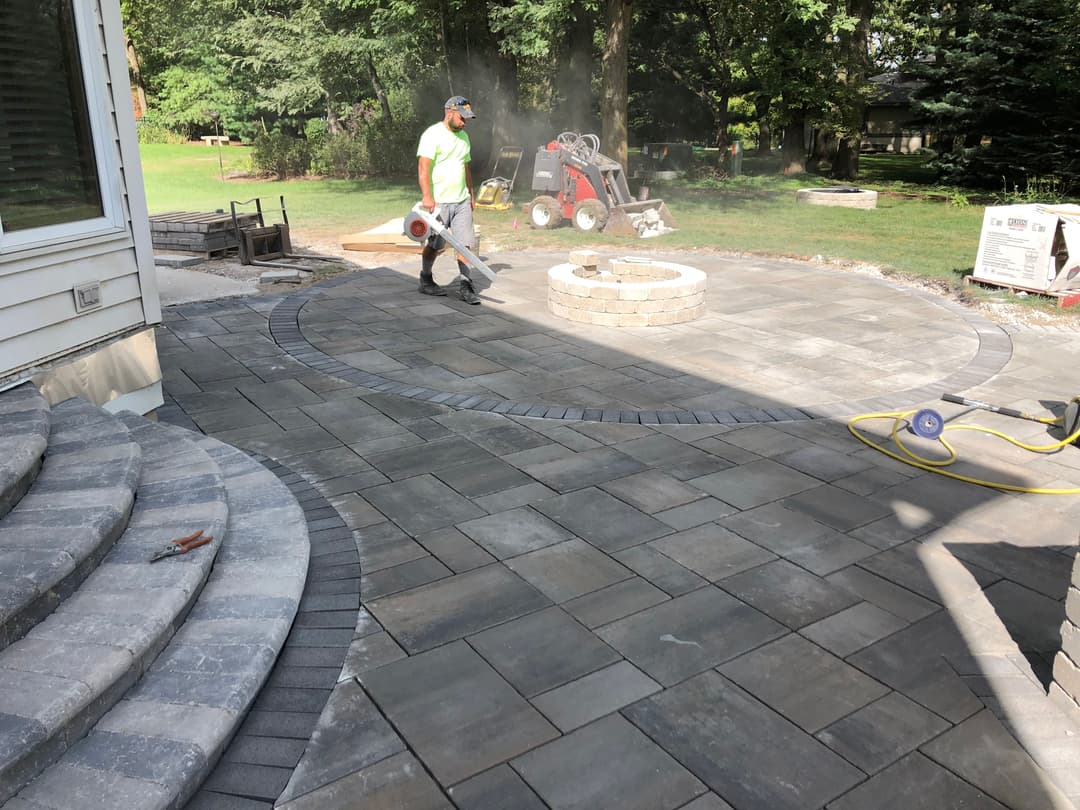 Worker using a leaf blower on a newly paved patio with a stone fire pit in a landscaped yard.