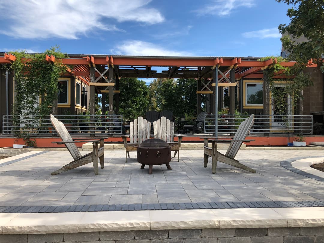 Outdoor seating area with Adirondack chairs and fire pit under a blue sky and greenery.